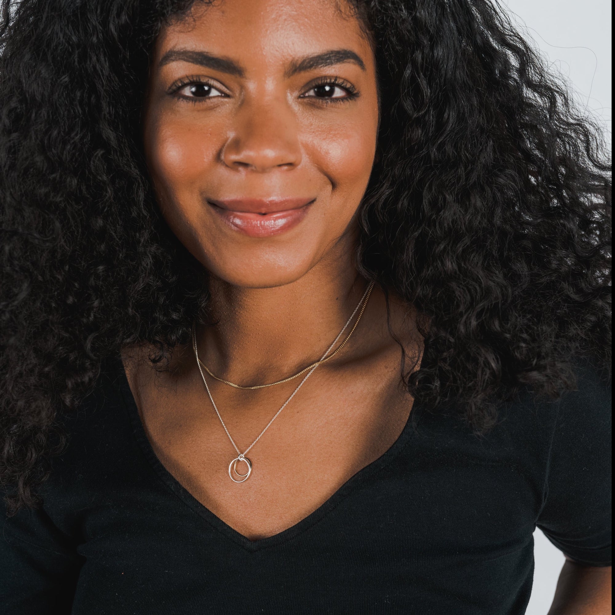 A person with long curly hair and a nose ring smiles slightly. They are wearing a black top and layered necklaces, one delicate with a circular pendant alongside the classic Curb Chain Necklace by Becoming Jewelry.