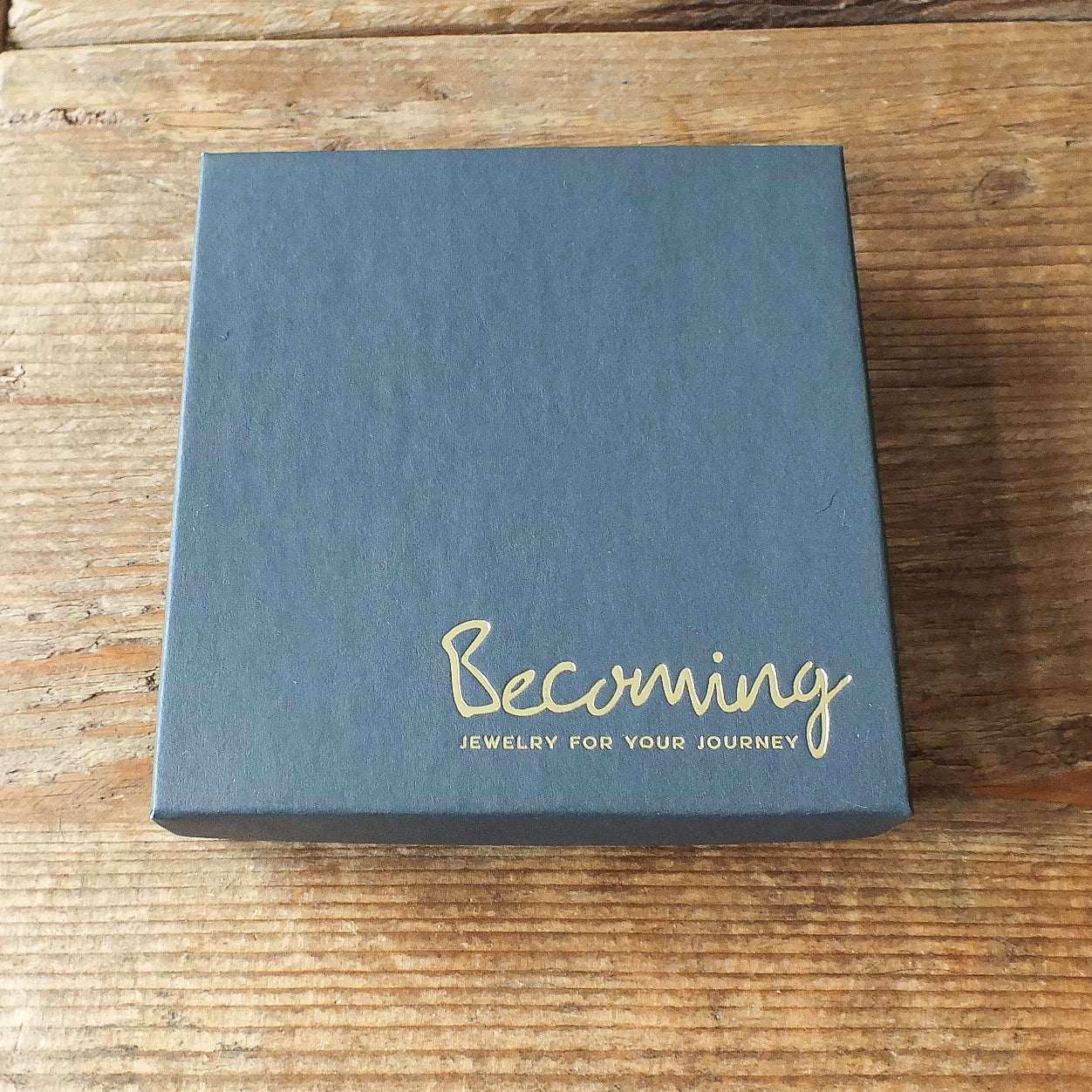 On a wooden surface lies a square, dark gray box adorned with the gold-etched Becoming Jewelry for Your Journey, carrying an aura of love and mindfulness, housing the May You Be Happy Necklace by Becoming Jewelry.