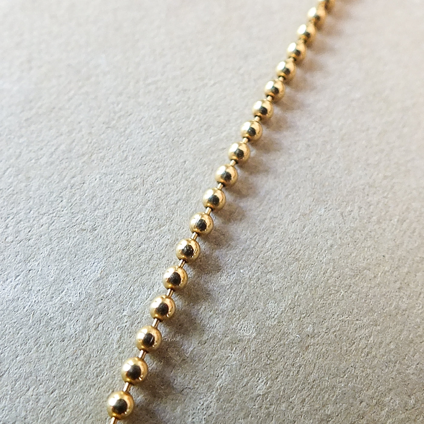 Close-up image of a gold Bead Chain Necklace by Becoming Jewelry, each bead connected in a linear sequence, layered gracefully on a textured beige surface.