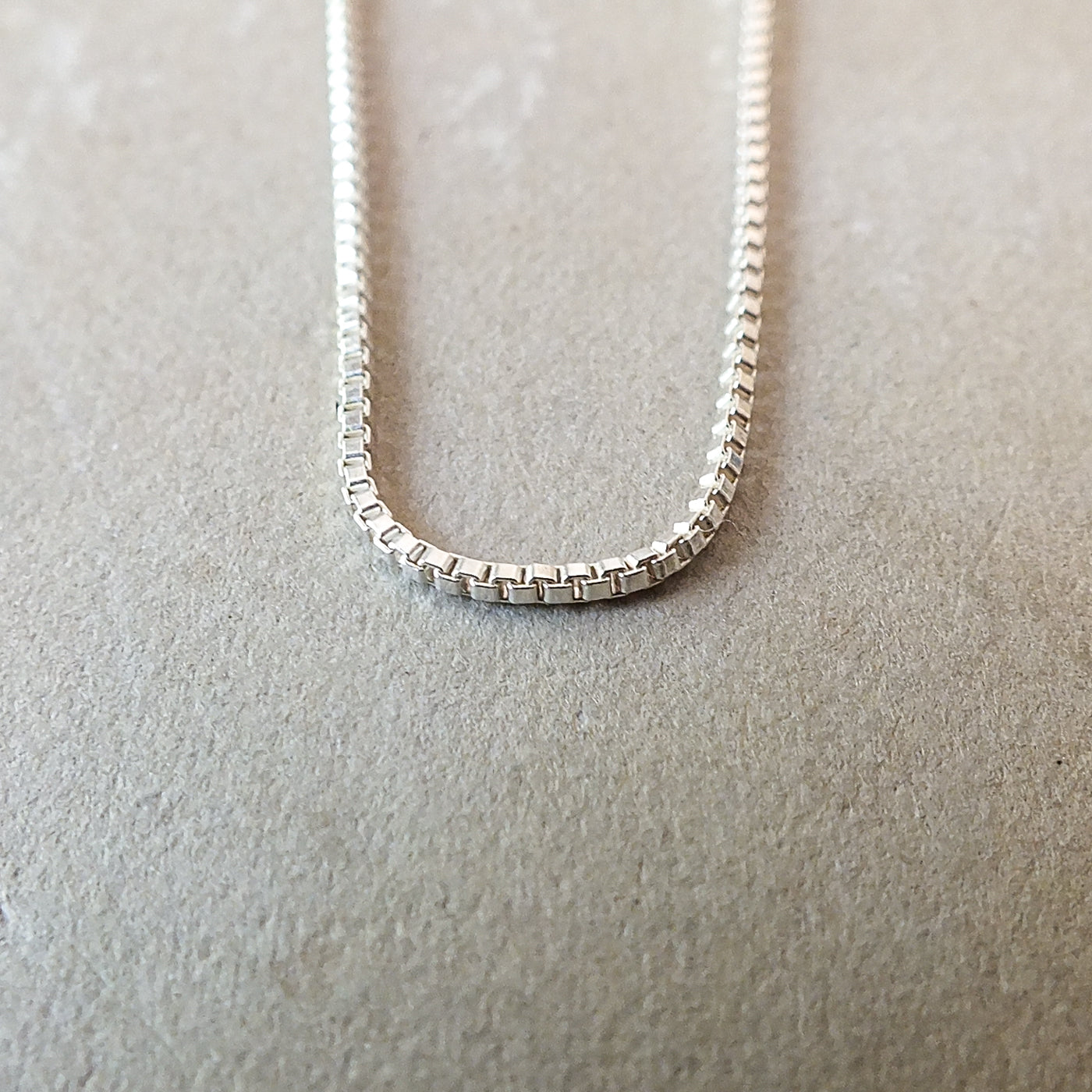 Close-up of the lightweight Becoming Jewelry Box Chain Necklace with small rectangular links, displayed on a light beige surface, exuding a classic look.