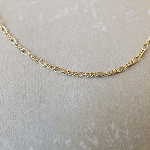 A gold-filled Becoming Jewelry Figaro Chain Necklace laid straight on a textured surface.
