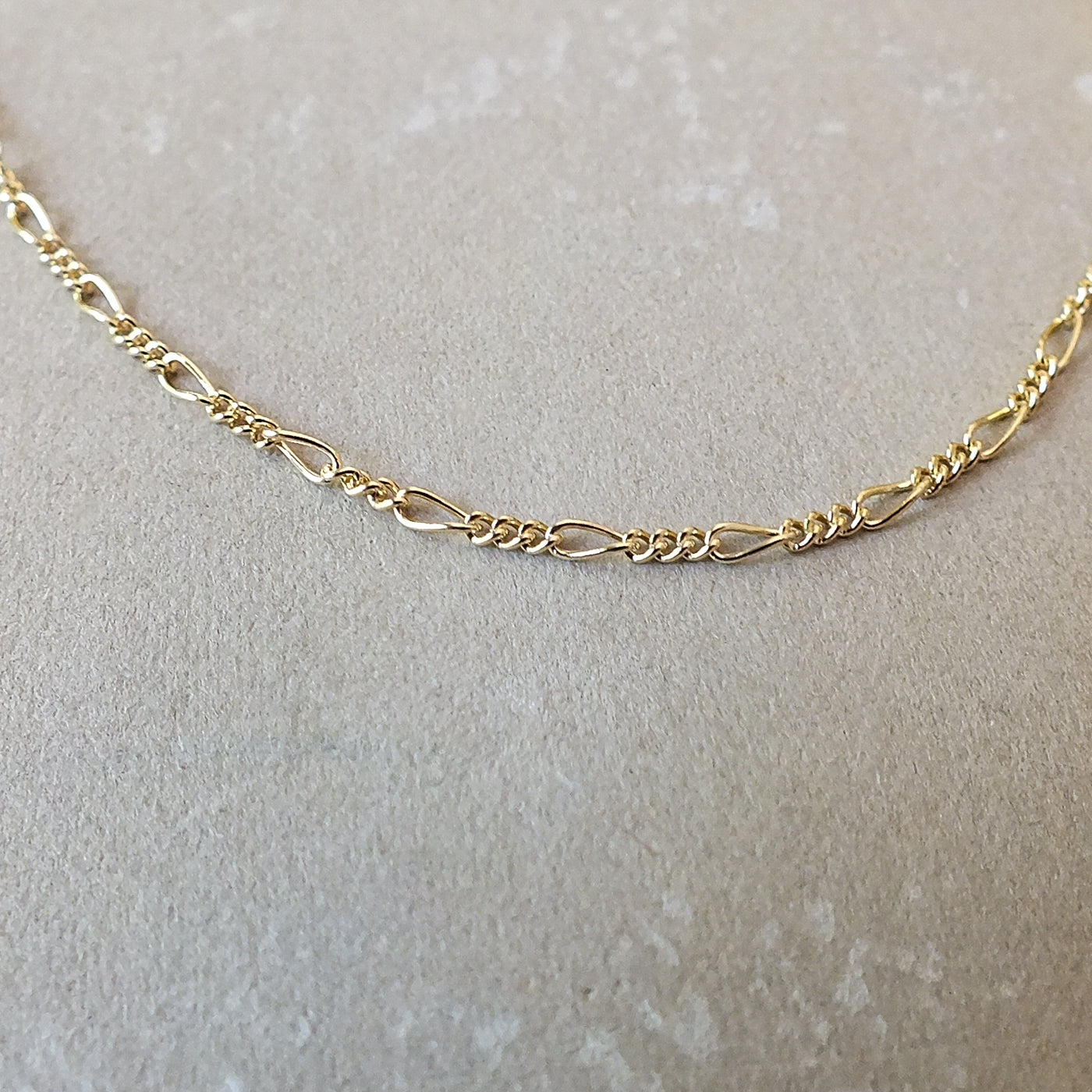 A gold-filled Becoming Jewelry Figaro Chain Necklace laid straight on a textured surface.