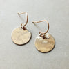 A pair of Becoming Jewelry Hammered Disc Drop Earrings, small with hook backings displayed on a grey surface.