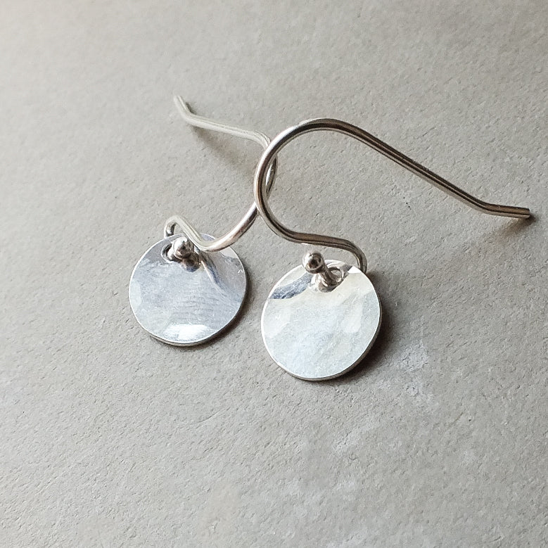 A pair of tiny Becoming Jewelry Hammered Disc Drop Earrings on a gray surface.