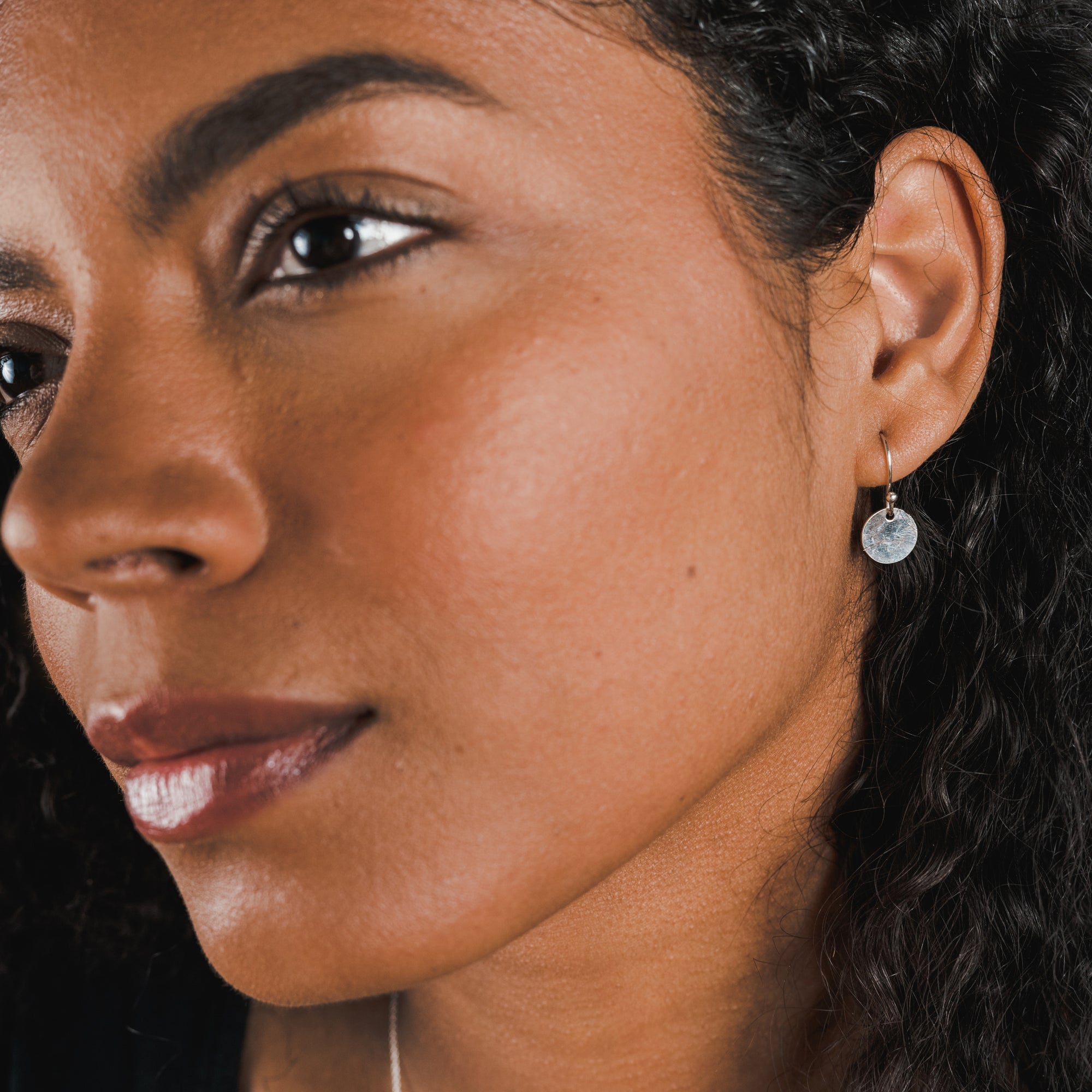 Close-up of a person with dark curly hair, wearing a tiny Hammered Disc Drop Earring by Becoming Jewelry on the left ear, looking off to the side—perfect for everyday wear.