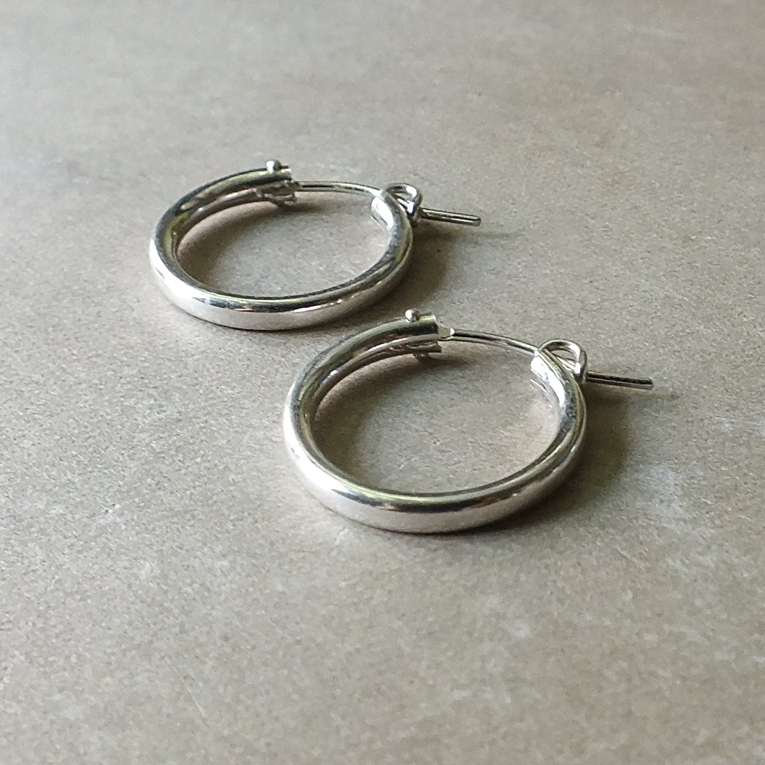 Close-up of a pair of large, classic silver hoop earrings by Becoming Jewelry laying on a gray surface—perfect for everyday wear.