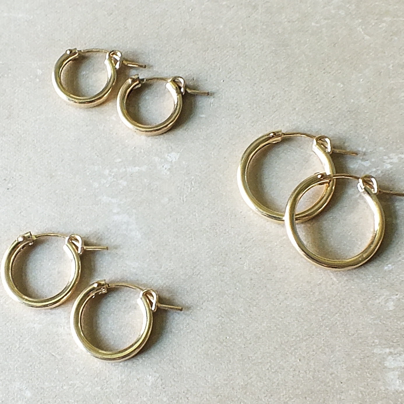 A set of four pairs of Becoming Jewelry&#39;s Everyday Hoop Earrings in classic gold, varying sizes, and perfect as lightweight accessories for any day, elegantly displayed against a light beige surface.