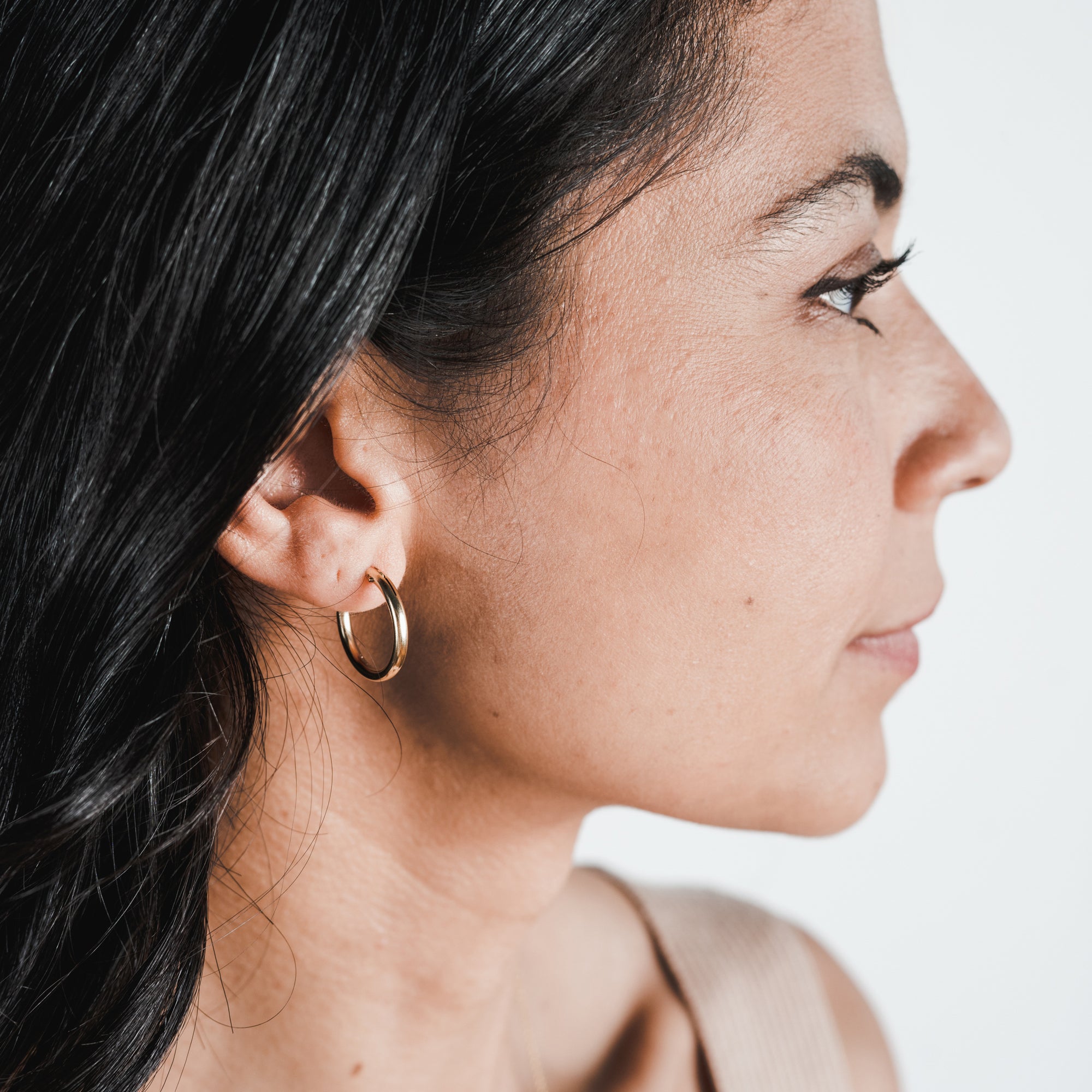 Profile of a woman with dark hair, wearing the large &quot;Everyday Hoop Earrings&quot; by Becoming Jewelry, looking to the right against a plain background.