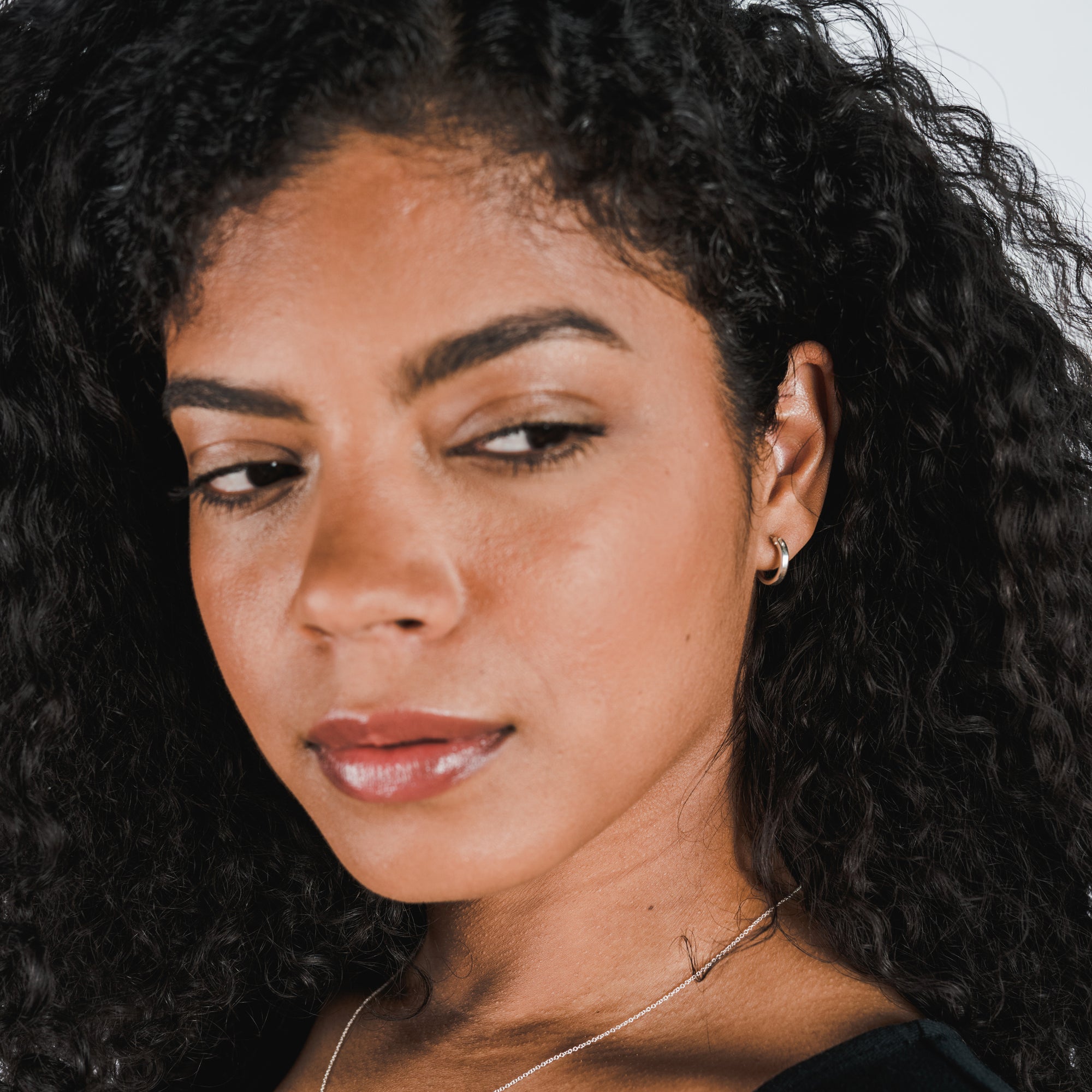A close-up of a person with curly dark hair, wearing Becoming Jewelry&#39;s Everyday Hoop Earrings (small) and a necklace, looking slightly to the side with a neutral expression.