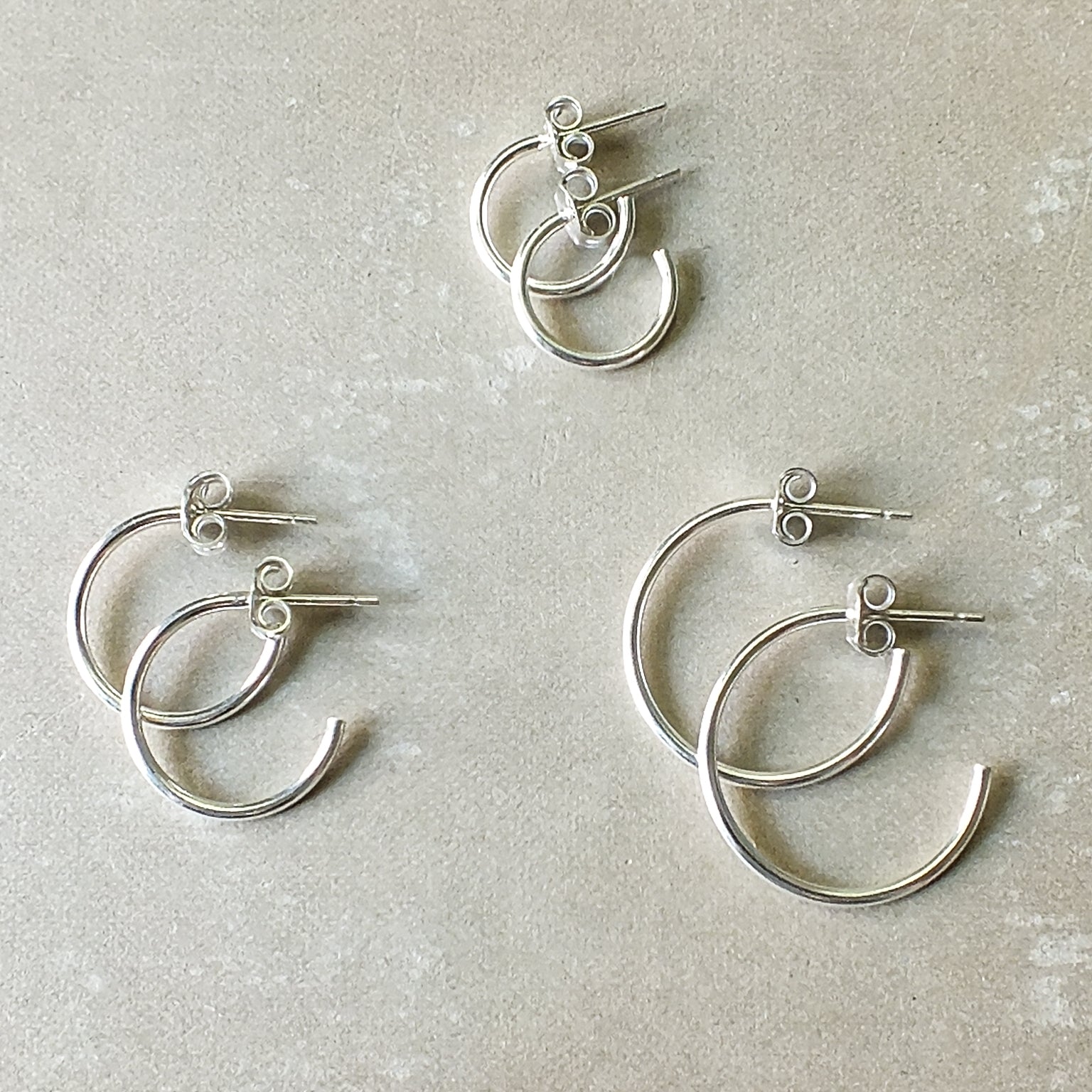 Four pairs of Becoming Jewelry&#39;s Open Hoop Earrings in a classic silver finish and varying sizes are arranged on a light gray surface, showcasing a versatile assortment of lightweight hoops.