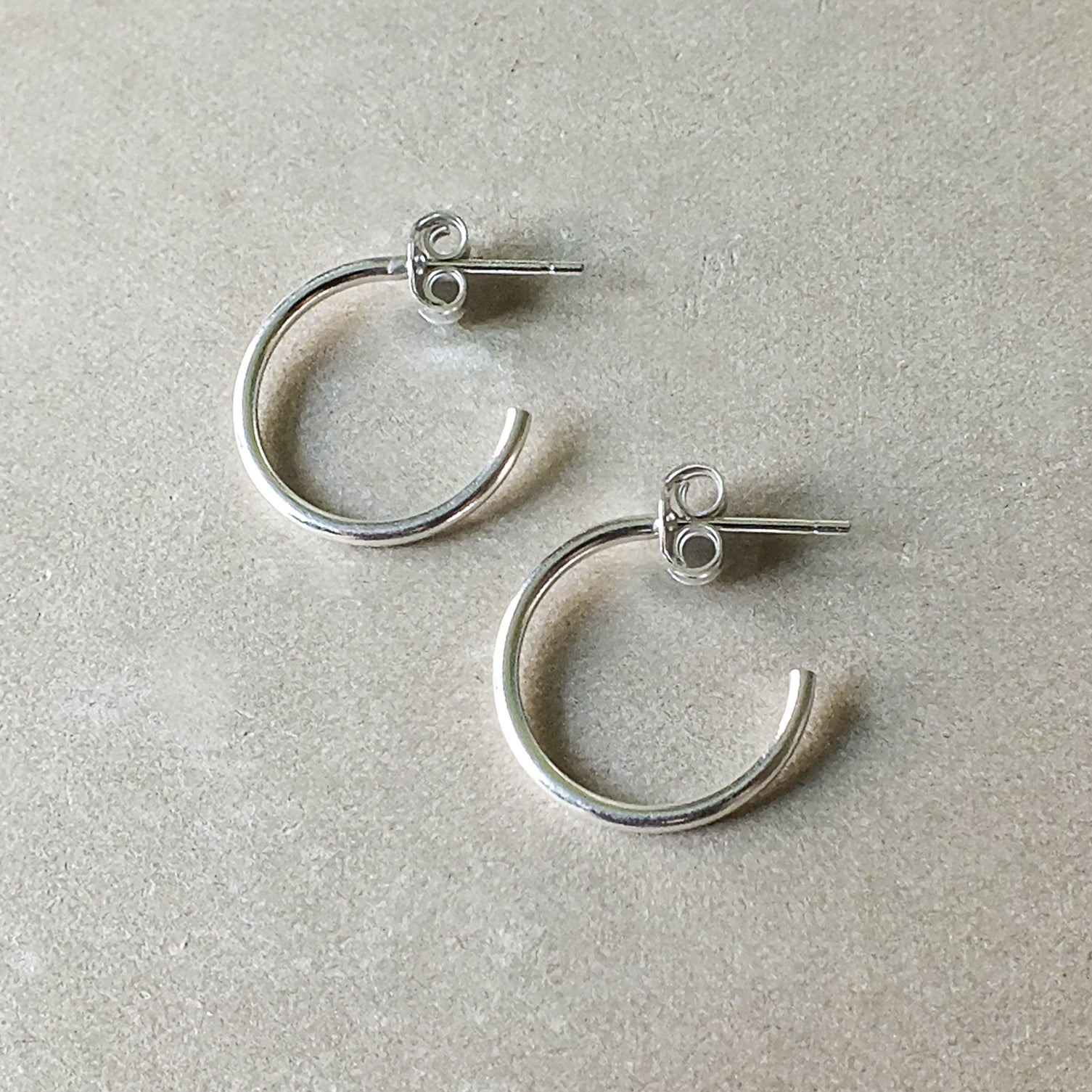 A pair of medium Open Hoop Earrings by Becoming Jewelry, lying on a textured beige surface, embodying lightweight classic elegance.