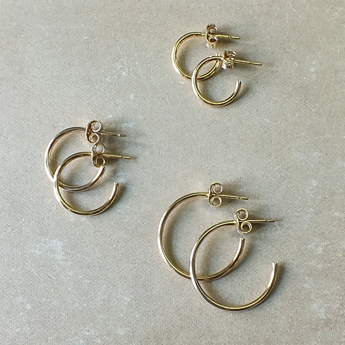 Three pairs of classic gold hoop earrings from Becoming Jewelry, including their Open Hoop Earrings in medium size, are displayed on a plain surface. These dainty hoops are lightweight, making them perfect for everyday wear.