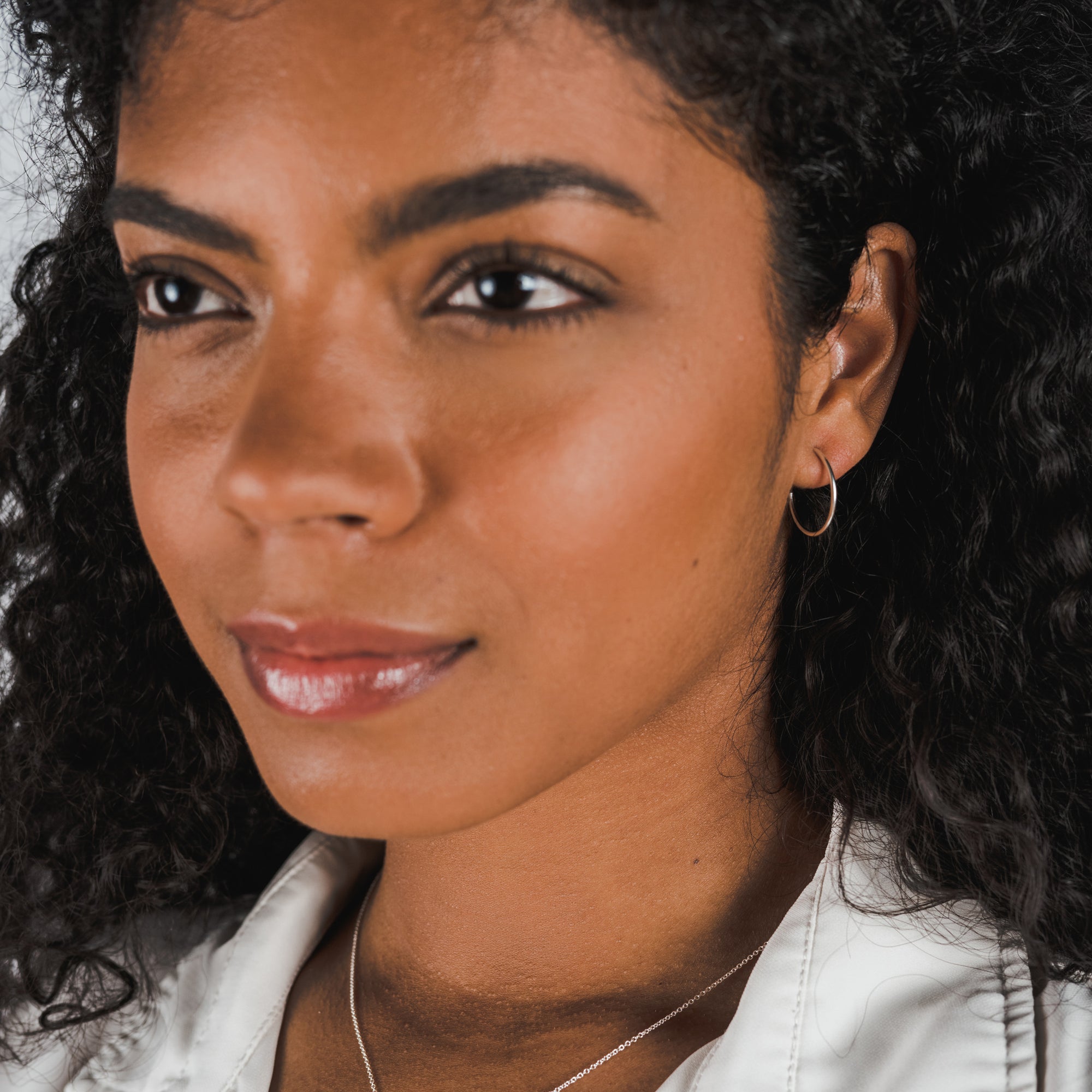 Close-up of a woman with curly hair wearing Becoming Jewelry&#39;s large Open Hoop Earrings, a dainty necklace, and a white top, looking slightly to the right.