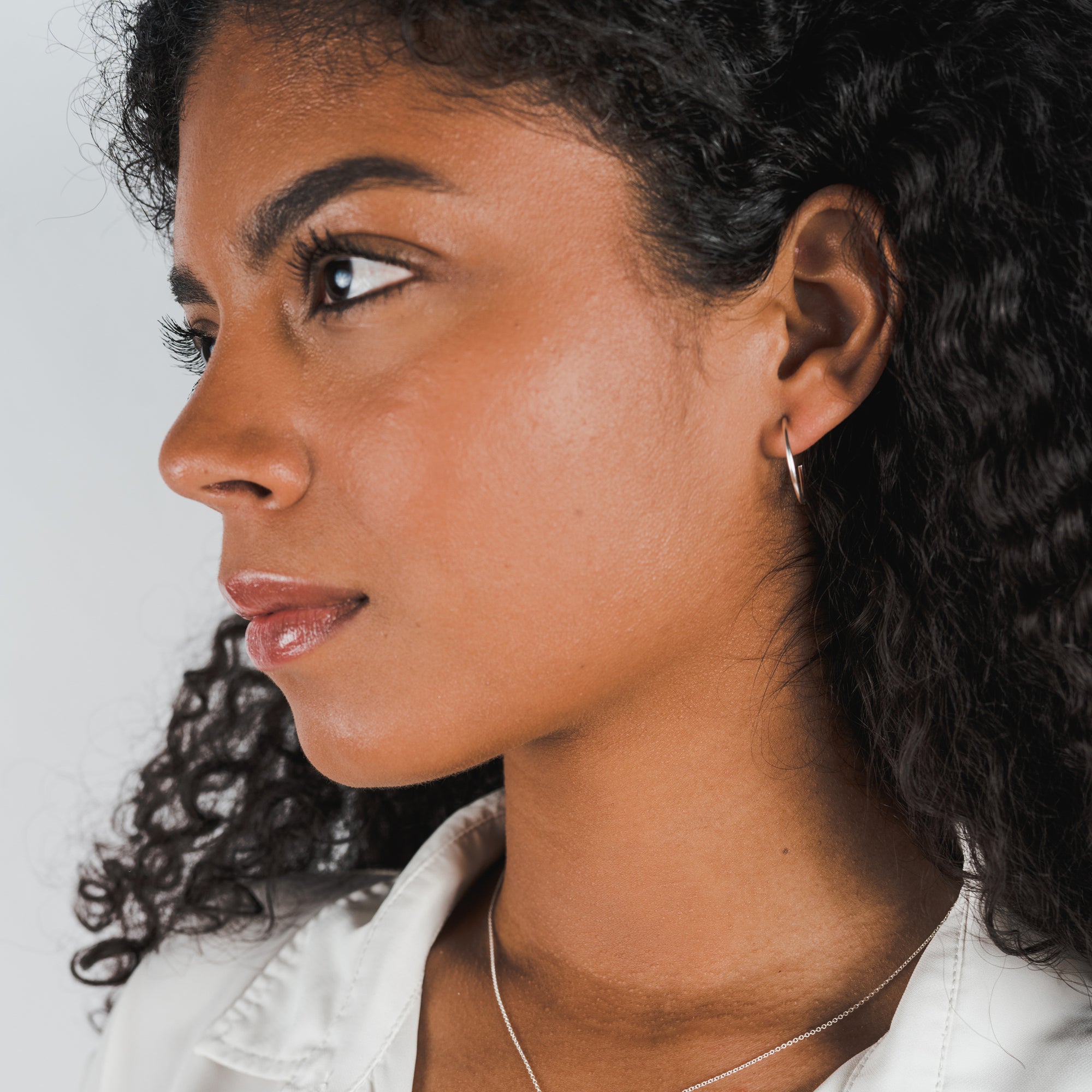 Profile view of a person with curly hair, wearing a white shirt, large Open Hoop Earrings by Becoming Jewelry, and a dainty necklace, looking towards the right.