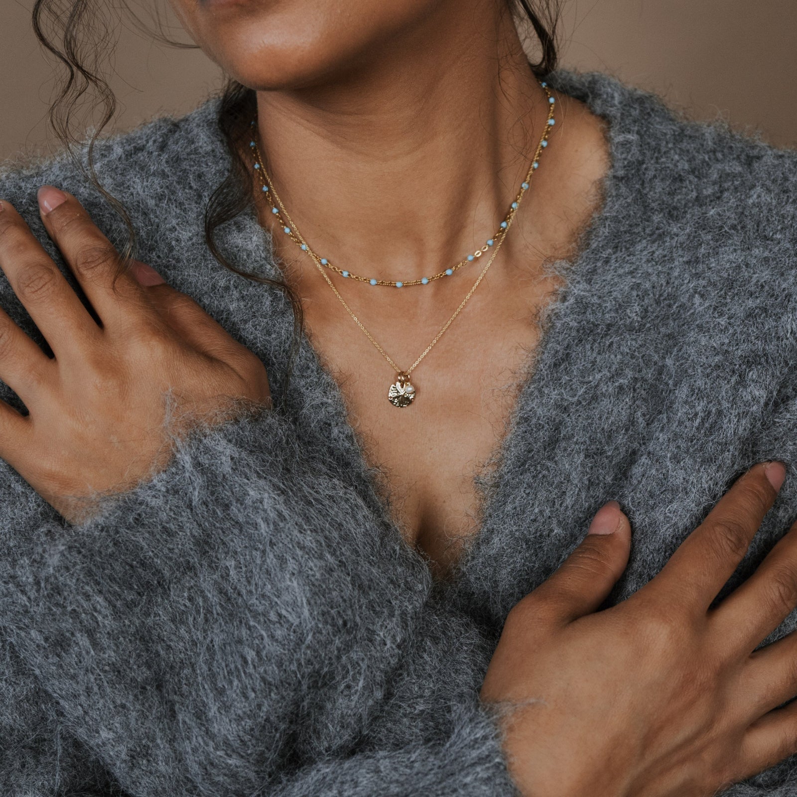 Person wearing a gray sweater with layered necklaces against a brown background