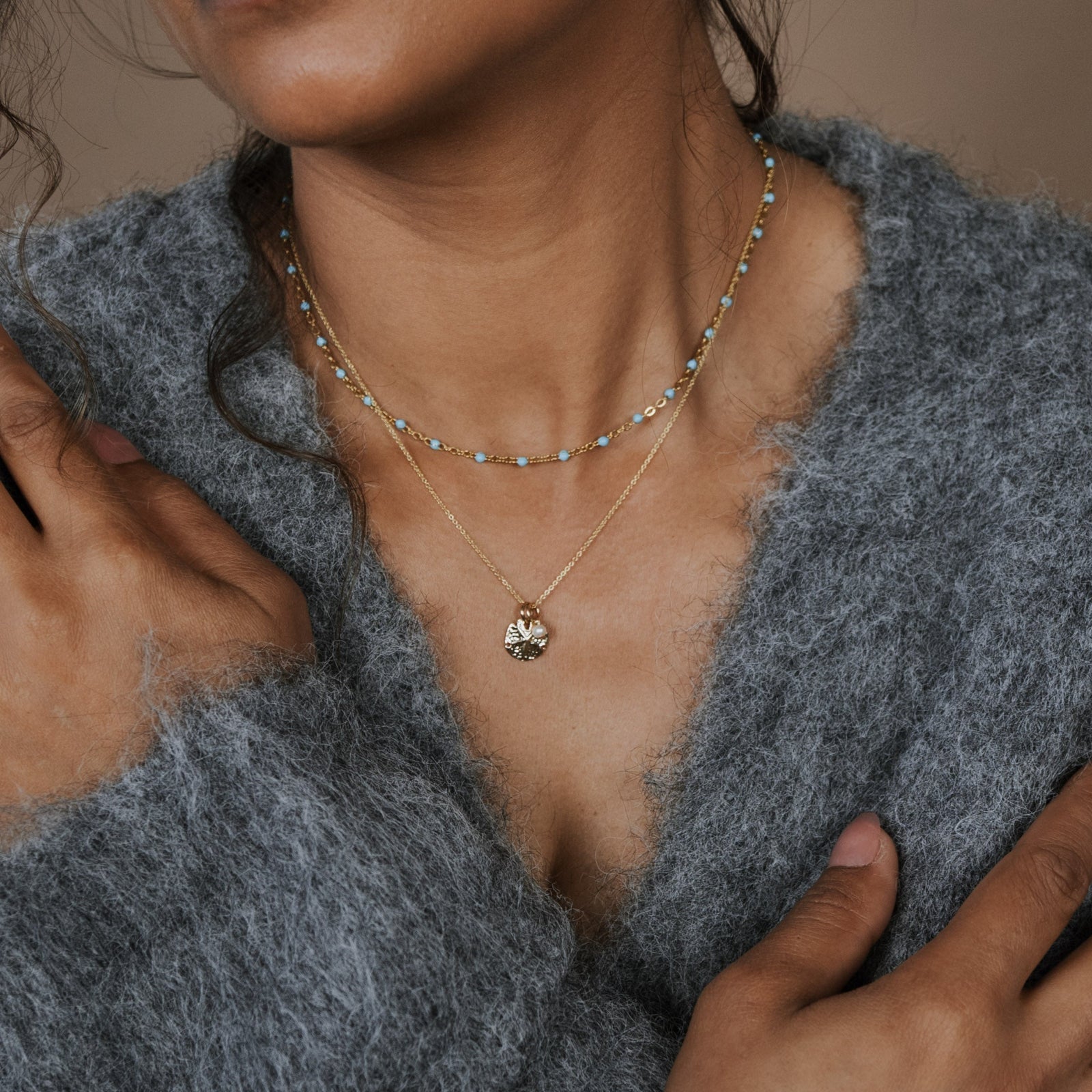 Person wearing a gray sweater with layered necklaces against a brown background