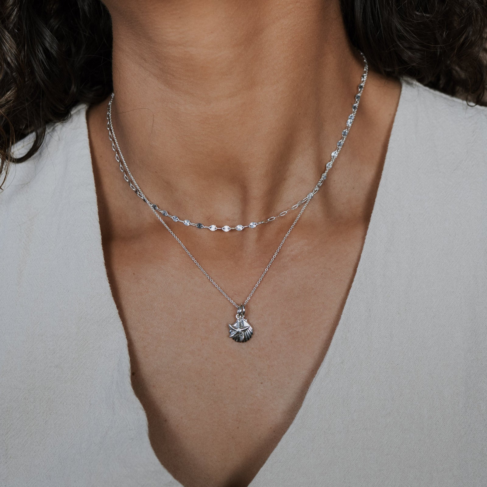 Close-up of a person wearing a silver necklace with a pendant against a neutral background