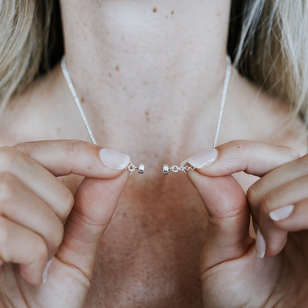 A person wearing a silver Stick Together Necklace by Becoming Jewelry is delicately clasping it near their neck, emphasizing its unique connection and magnetic beads.