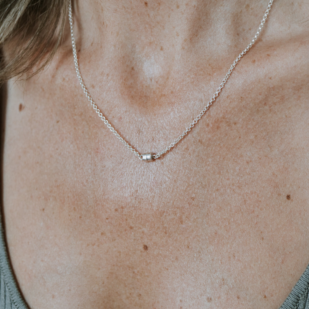 A person sporting a Becoming Jewelry Stick Together Necklace, featuring a small pendant on a silver chain, over a gray top, symbolizing a special connection.