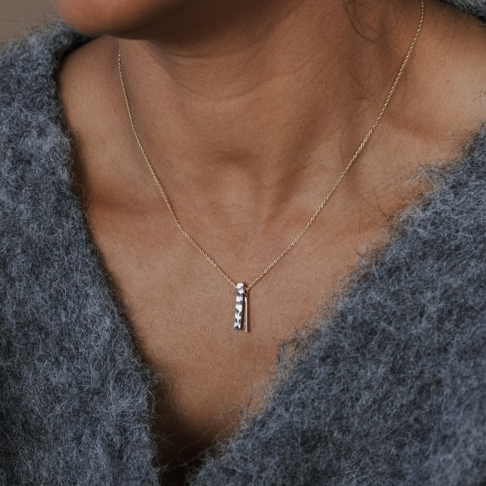 A close-up of a person wearing a delicate gold chain necklace with a vertical bar pendant set with small clear stones, paired with a fuzzy gray knit top.