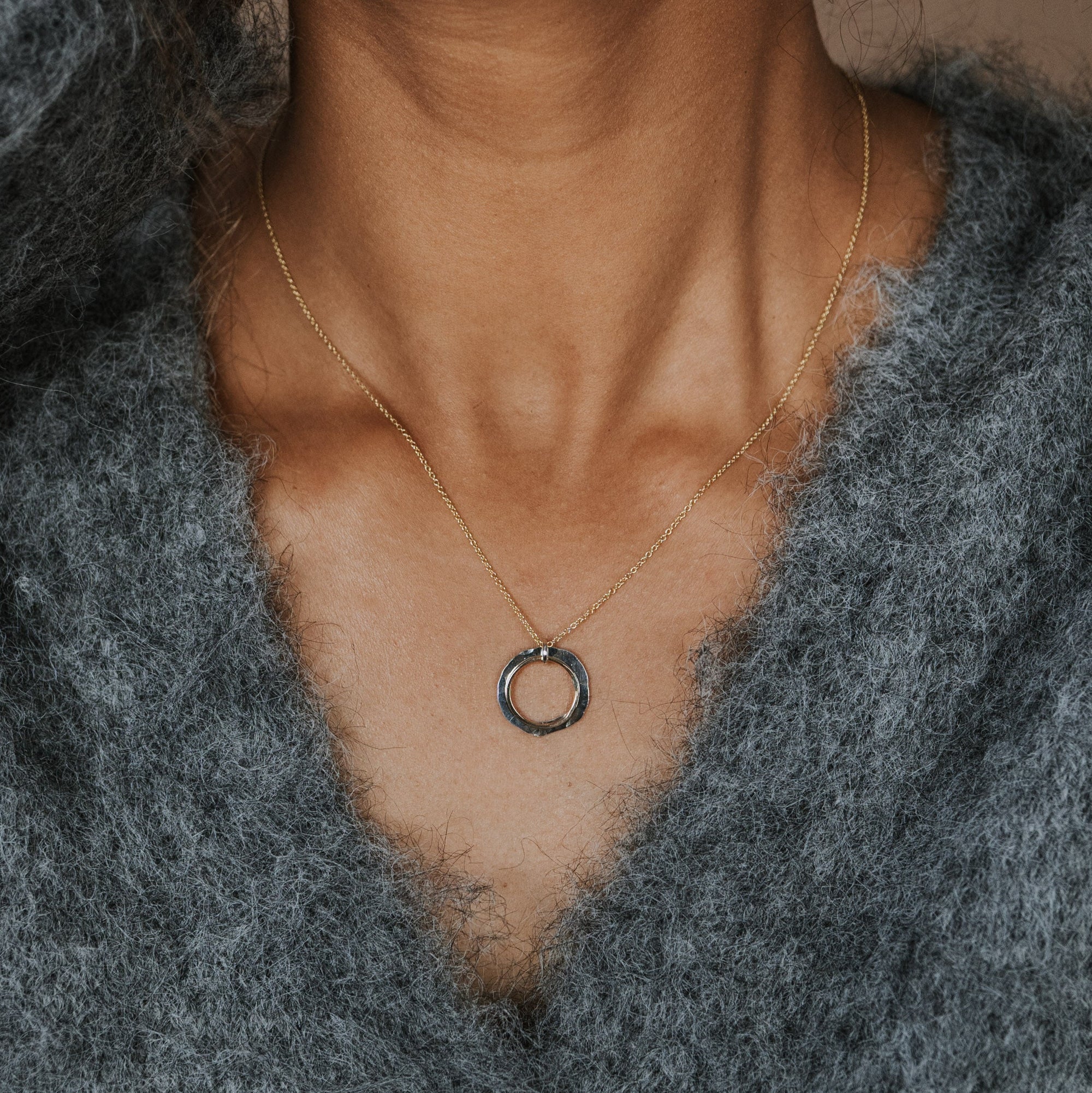 A close-up of a person wearing a delicate gold chain necklace with a small circular ring pendant, styled with a fuzzy gray button-up sweater.