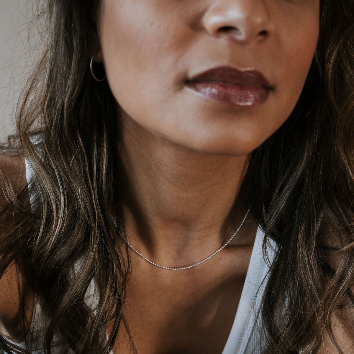 A woman in a white tank top layers her wavy hair over her shoulders, accenting a glossed smile with hoop earrings and the delicate Bead Chain Necklace from Becoming Jewelry.