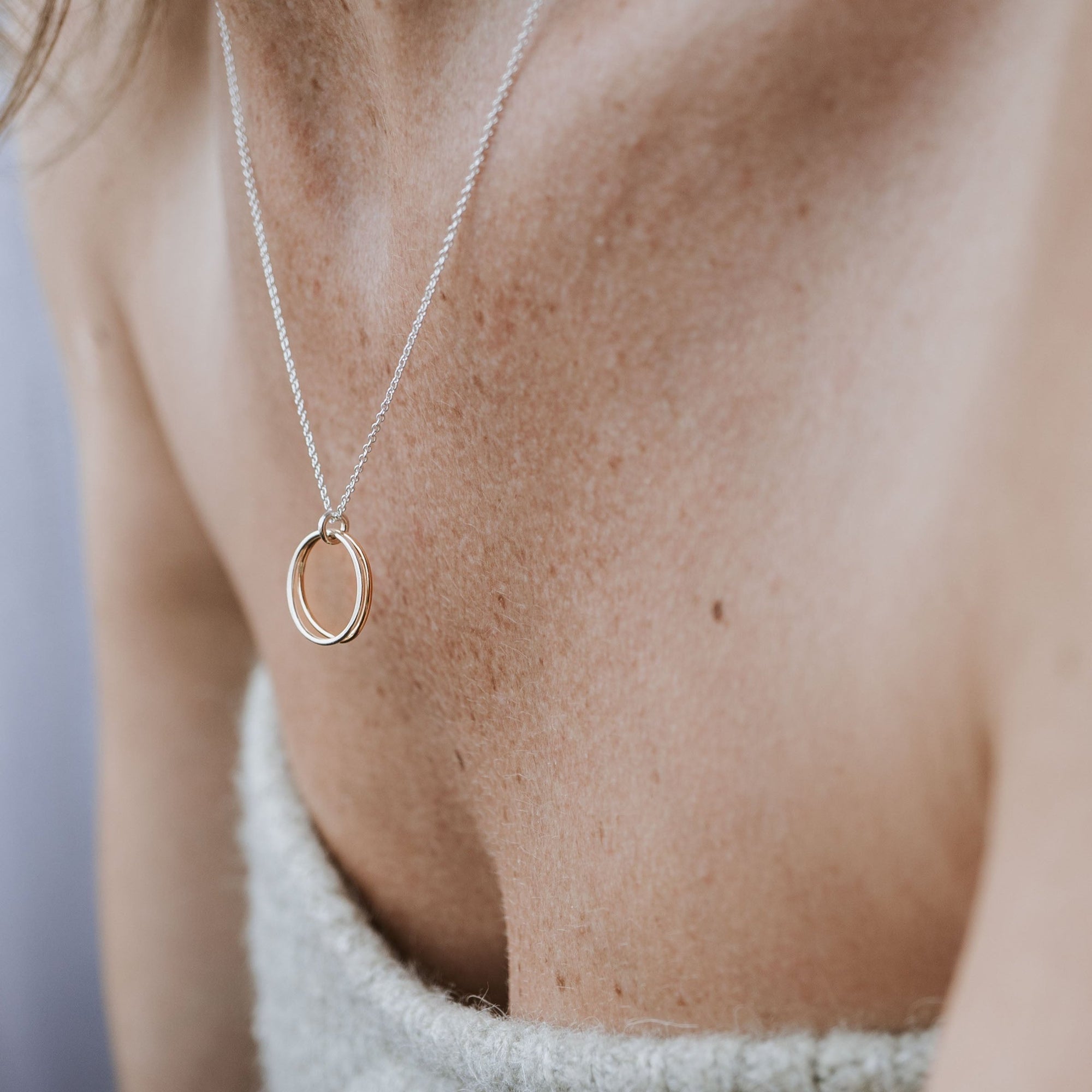 Close-up of a person wearing the Becoming Jewelry Friendship Circles Necklace, a perfect gift. The mixed metal double-ring pendant rests elegantly against bare skin, complemented by a textured light-colored top.