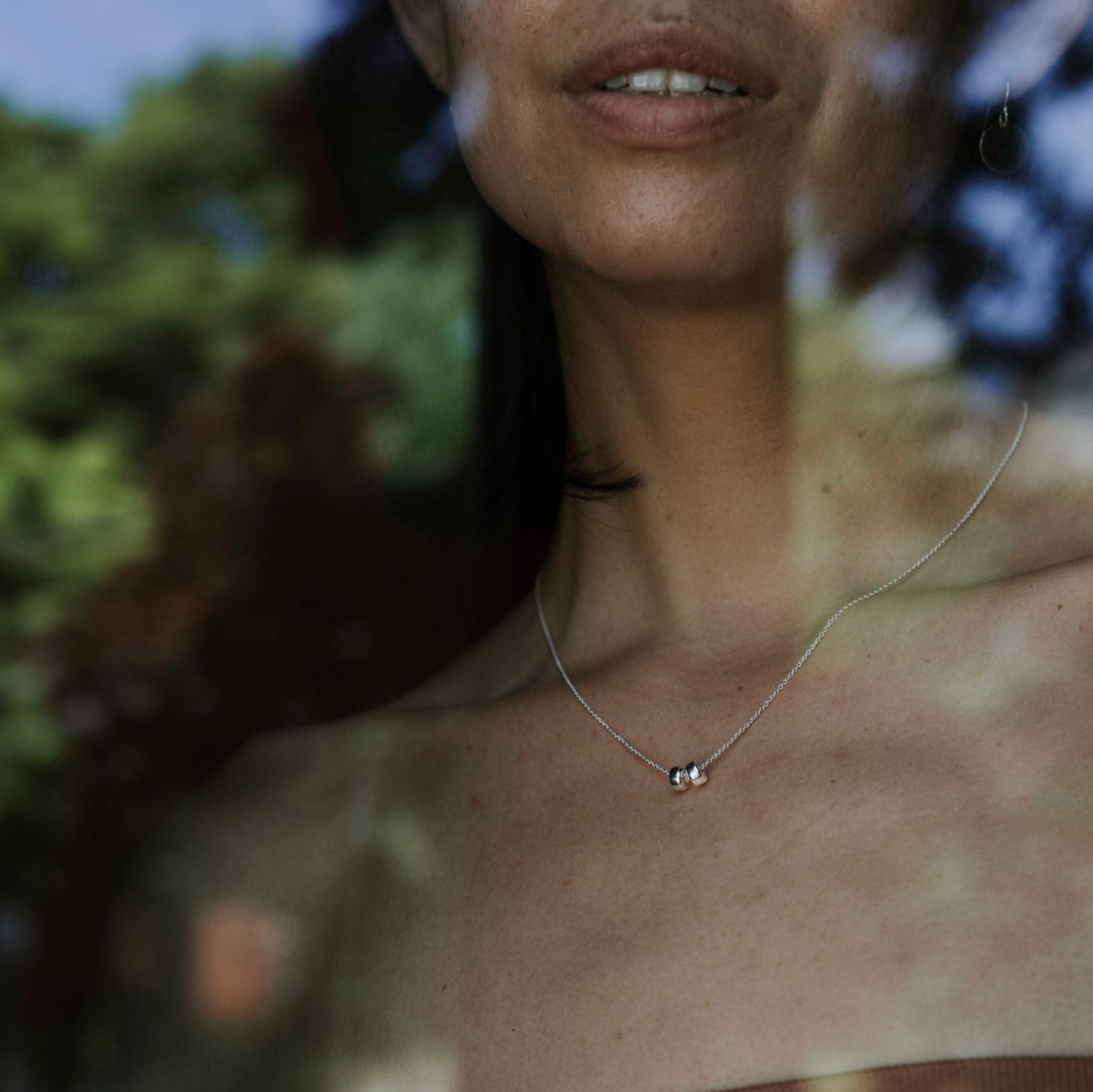 Close-up of a woman&#39;s neck and lower face, adorned with Becoming Jewelry&#39;s Friends Beads Necklace—a delicate silver accessory featuring a pendant shaped like two intricately intertwined circles, symbolizing the deep bond between dearest friends—set against a blurry, leafy background.