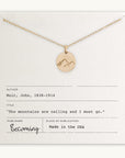 Becoming Jewelry's Mountains Are Calling Necklace pendant necklace with mountain charm displayed on a card with bibliographic-style information, including a quote, "the mountains are calling and I must go.