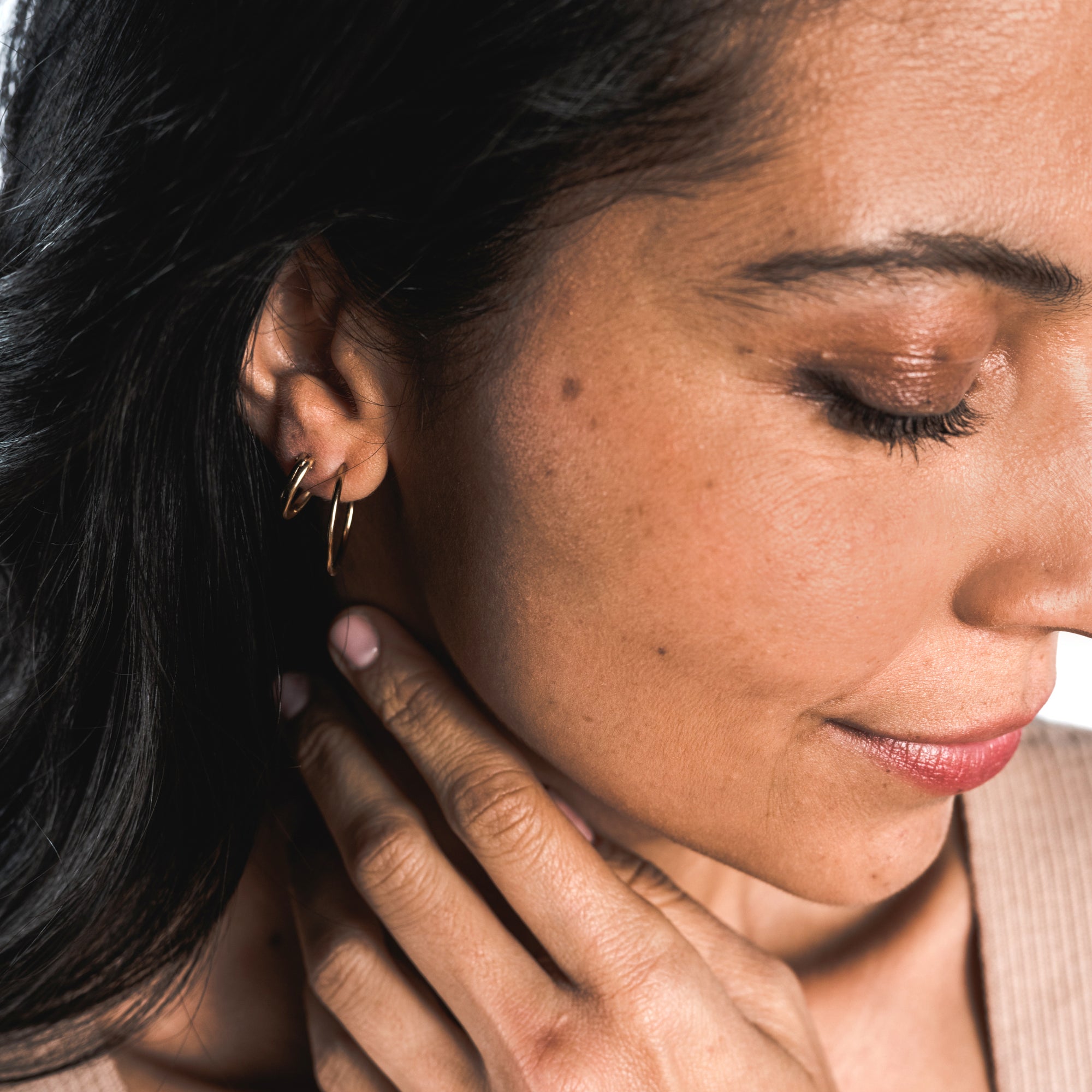 A person with long, dark hair wearing Becoming Jewelry&#39;s large Open Hoop Earrings and a beige top, gently touching their neck with their hand.