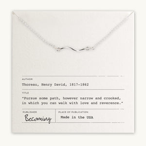 A Path Necklace with a curved path charm in gold filled displayed on a card featuring a quote by Henry David Thoreau, with the mention "becoming" and "made in the USA. (by Becoming Jewelry)