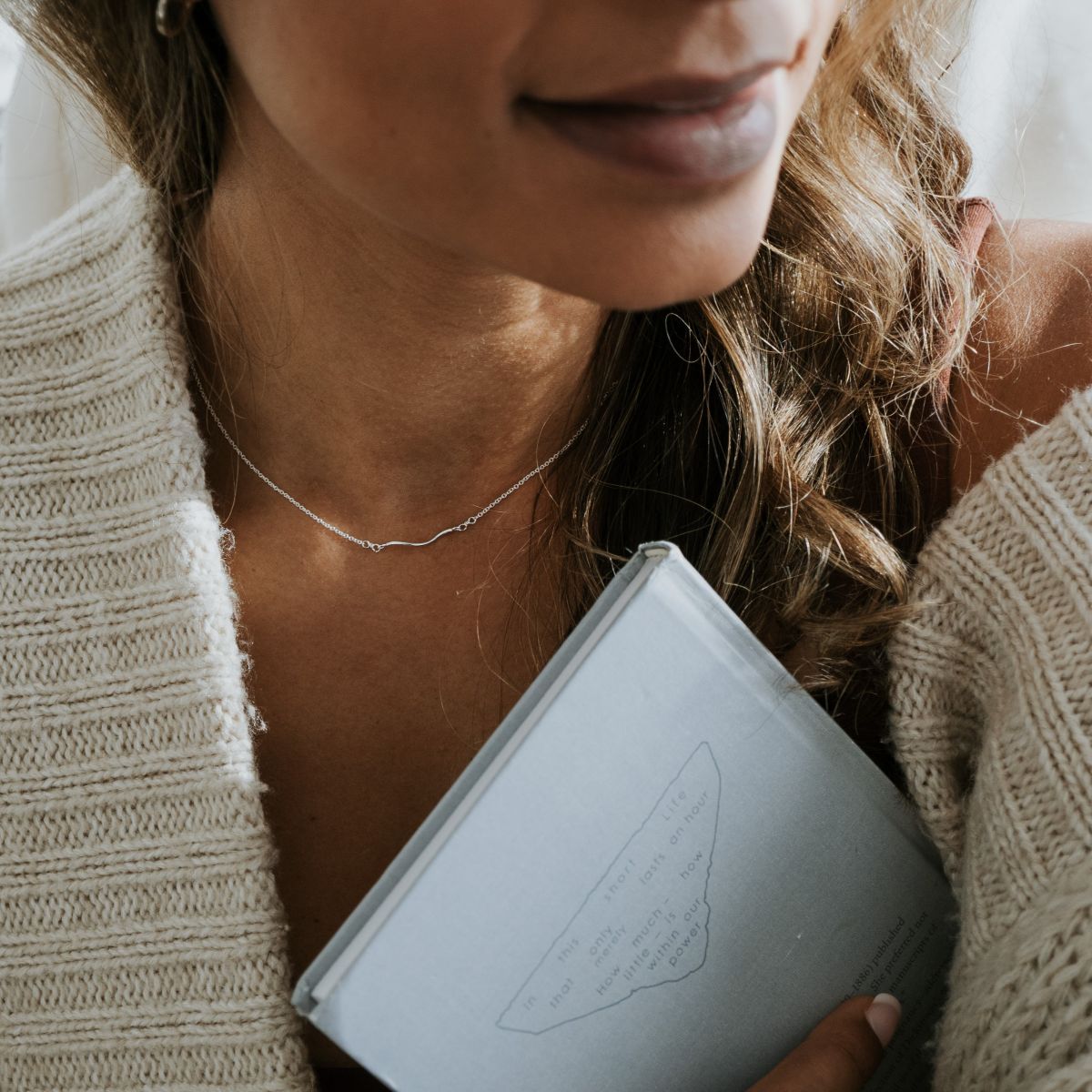 A person clutches a blue book close to their heart, wearing a knitted sweater and the Path Necklace by Becoming Jewelry, with long, wavy hair partially visible.