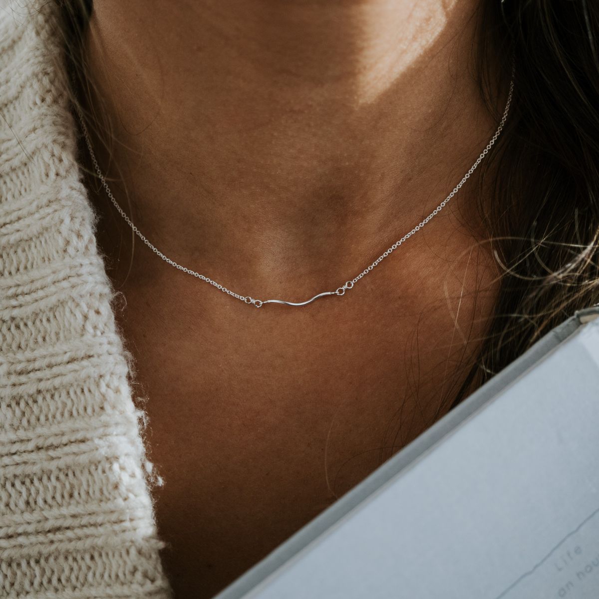 Close-up of a person wearing the Becoming Jewelry Path Necklace, a silver piece with a heart-like curve design, paired with a knitted sweater while holding a book.