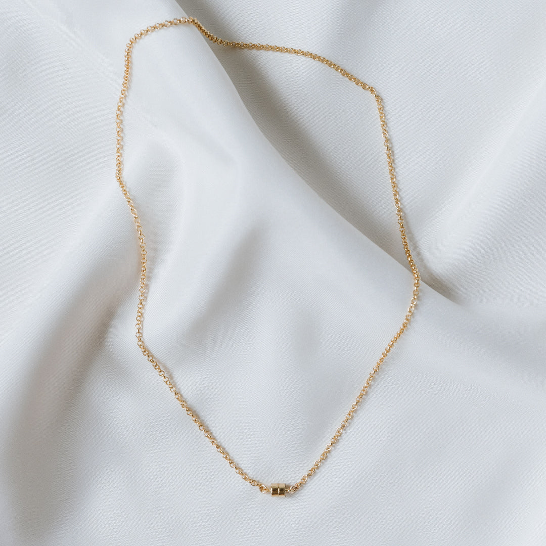 A charming Stick Together Necklace by Becoming Jewelry, showcasing a gold chain with a petite rectangular pendant, elegantly laid out on a white fabric background to represent a meaningful bond.