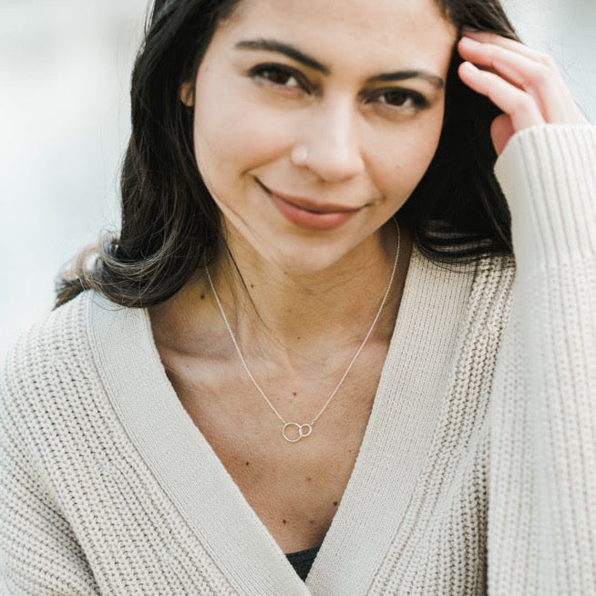 A woman with long dark hair wears a beige knit cardigan and a necklace adorned with intertwined circles. She smiles slightly while touching her hair, the Sisters Necklace—a gift from her sister by Becoming Jewelry—gleaming softly in the light.