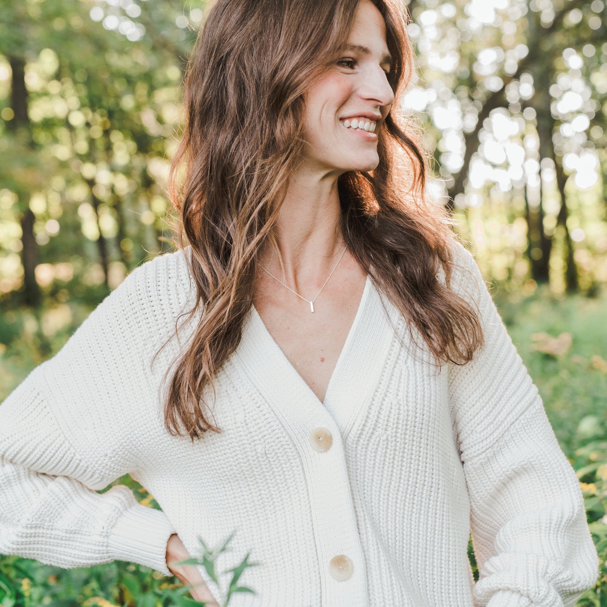 A woman with long, wavy brown hair wearing the Pillar of Strength Necklace by Becoming Jewelry stands outdoors, smiling and looking to the side. The background is a forest with sunlight filtering through the trees, serving as a reminder of nature&#39;s strength.