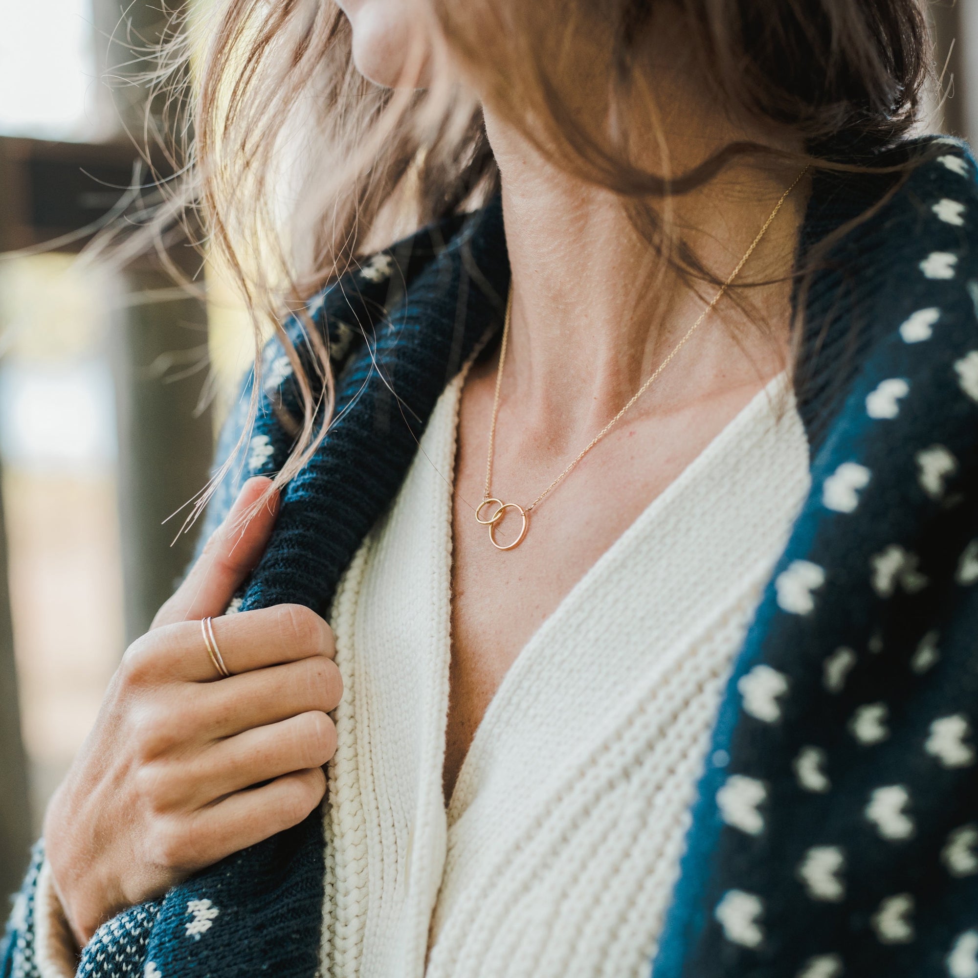 Close-up of a person wearing a white knitted top and a navy blue patterned cardigan, showcasing the Becoming Jewelry Mother Necklace with interlocking rings symbolizing mother&#39;s love, and a gold ring on their finger.