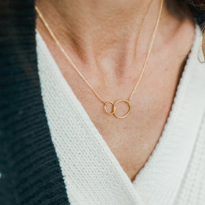 A woman wearing a white v-neck garment and a gold Becoming Jewelry mother necklace with two interlocking circles.