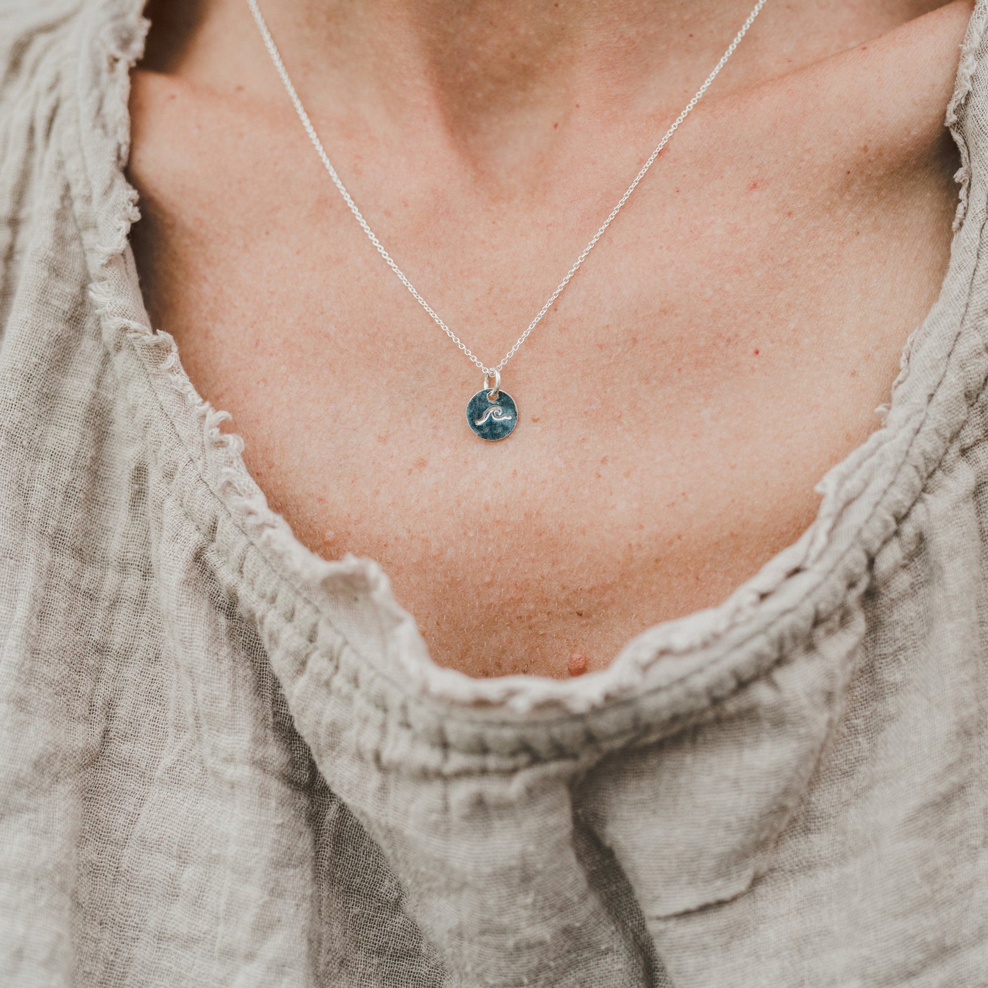 A close-up of a person&#39;s neck showing a Swim the Sea Necklace by Becoming Jewelry, with a wave charm on a sterling silver chain, partially framed by a beige, textured neckline of a garment.