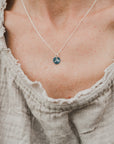 A close-up of a person's neck showing a Swim the Sea Necklace by Becoming Jewelry, with a wave charm on a sterling silver chain, partially framed by a beige, textured neckline of a garment.