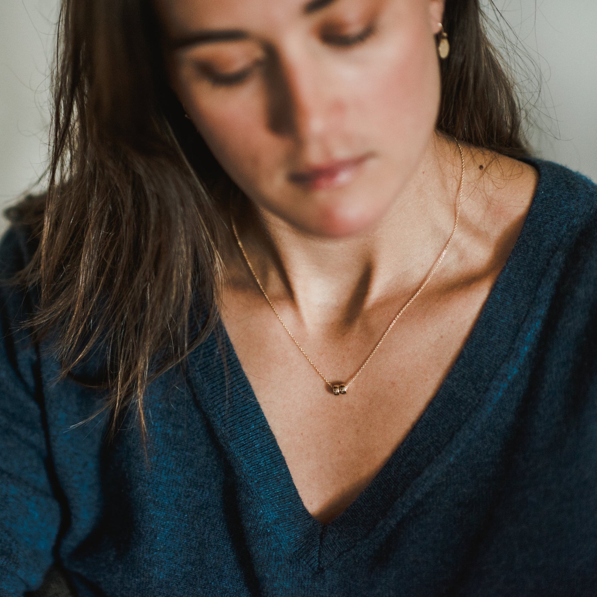 A woman with long brown hair, wearing a deep blue V-neck top and a &quot;Friends Beads Necklace&quot; from Becoming Jewelry—perhaps a meaningful gift from one of her dearest friends—looks down.
