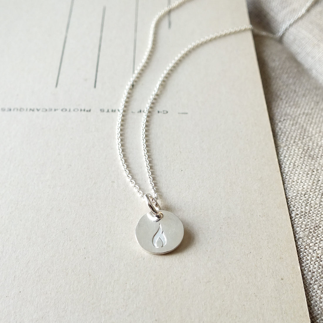 The On Fire Necklace by Becoming Jewelry, a silver accessory adorned with a round pendant engraved with a flame symbol, rests on a textured beige surface accented with faint lines and text in the background—an ideal gift for graduates or anyone seeking inspiration.