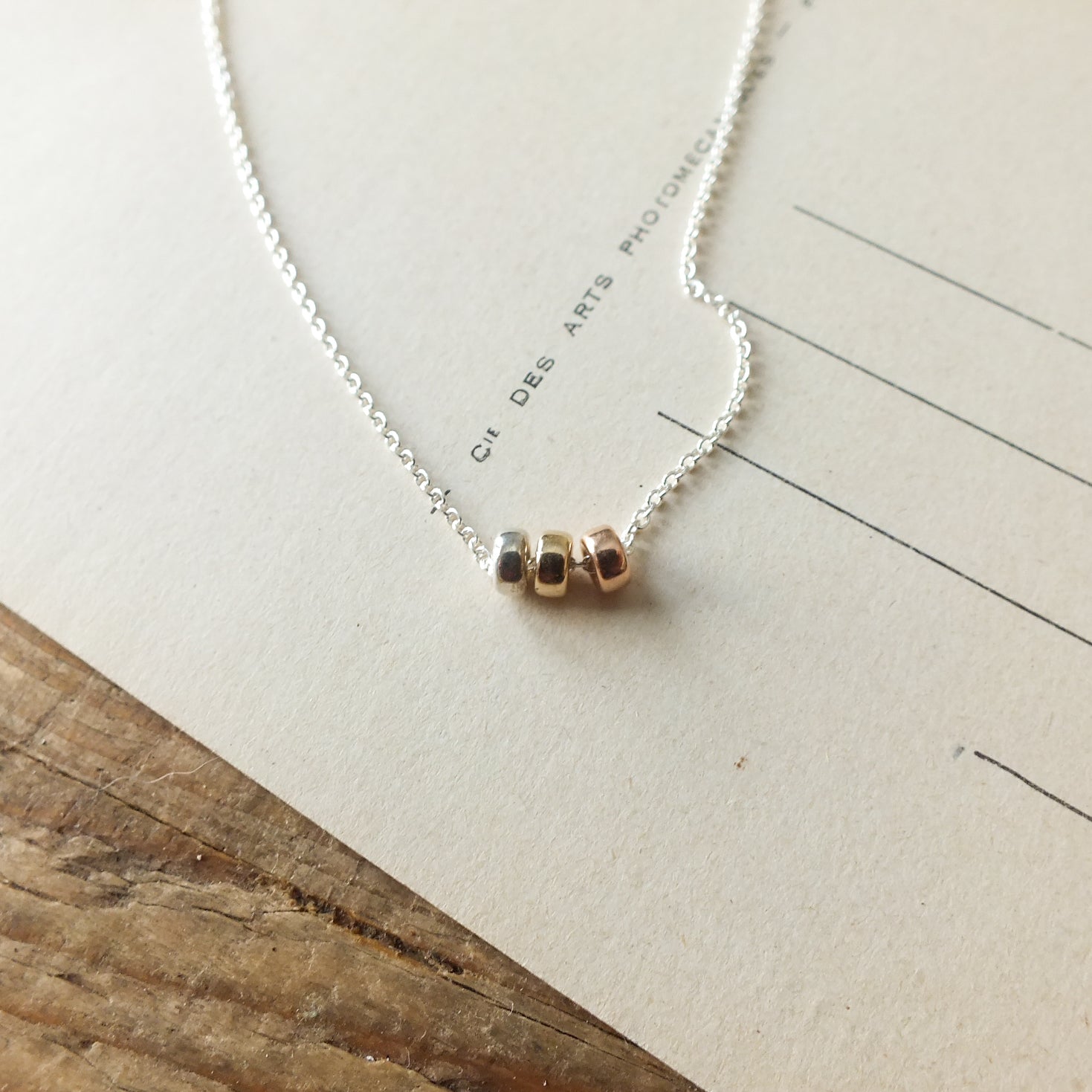 A delicate Three Things Necklace with three mixed metal beads displayed on a piece of paper by Becoming Jewelry.