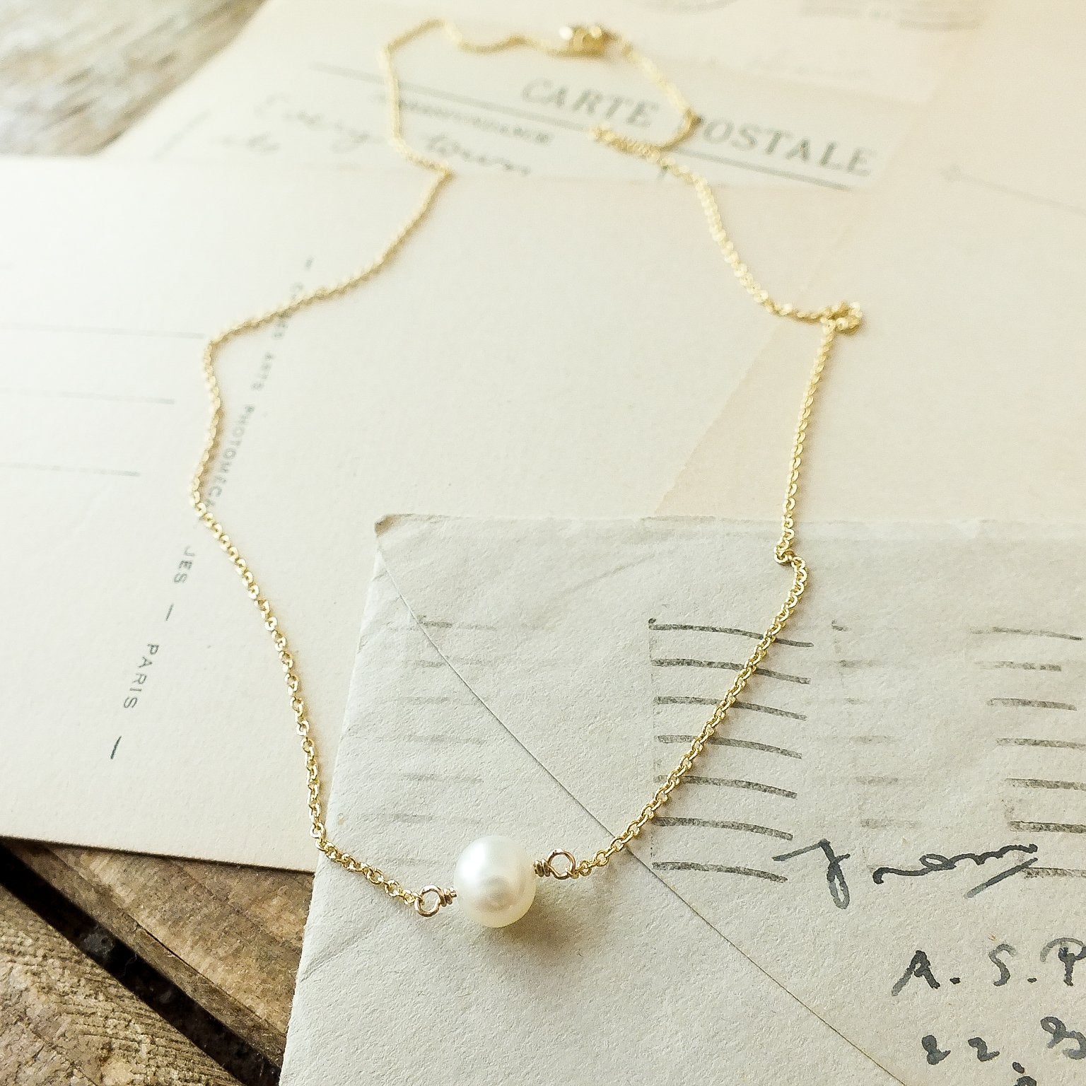 A delicate gold chain with a single pearl pendant, known as the Simple Pleasures Pearl Necklace by Becoming Jewelry, is laid on top of vintage postcards on a wooden surface.