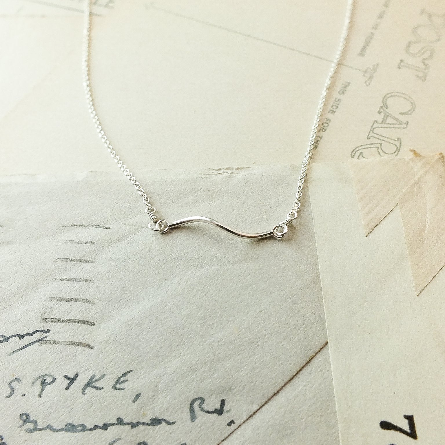 The Path Necklace from Becoming Jewelry: a delicate silver piece featuring a wavy pendant that gently reminds you of life&#39;s journey, beautifully showcased on vintage postcards.