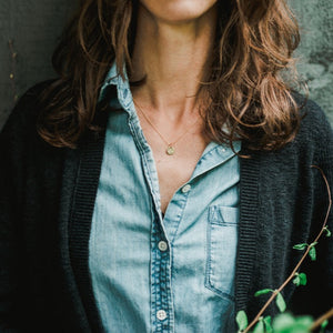Woman wearing a blue denim shirt under a black cardigan with a gold-filled You Are My Sunshine Necklace pendant by Becoming Jewelry, standing against a textured background.