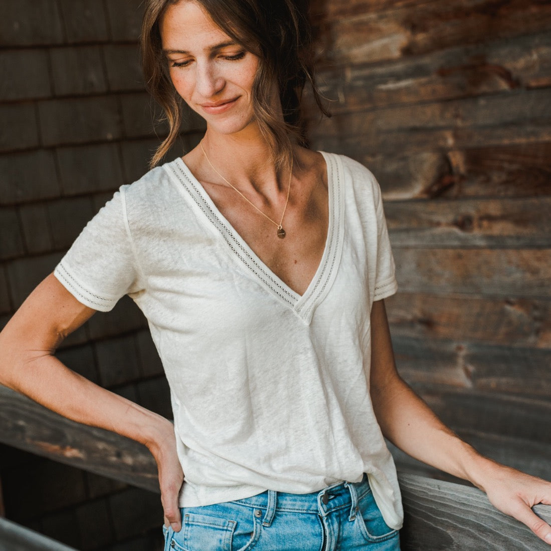 A woman in a casual white v-neck t-shirt and blue jeans leaning against a wooden wall, wearing a Becoming Jewelry Onward Necklace.