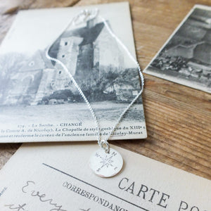 A Becoming Jewelry sterling silver Compass Necklace rests on a postcard featuring an old building, accompanied by other vintage correspondence on a wooden surface.
