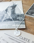 A Becoming Jewelry sterling silver Compass Necklace rests on a postcard featuring an old building, accompanied by other vintage correspondence on a wooden surface.