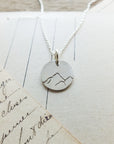 Sterling silver Mountains Are Calling necklace charm pendant on a necklace chain placed on handwritten paper by Becoming Jewelry.
